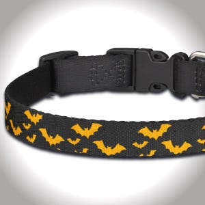 May include: A black pet collar featuring a repeating pattern of small orange bats. The collar is constructed from durable woven fabric and includes a black plastic side-release buckle, an adjustable slider, and a silver-toned metal D-ring attachment.