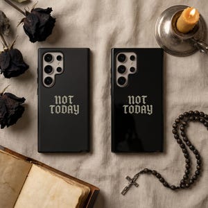 May include: Two black phone cases with the text "NOT TODAY" in a gothic font. The cases are on a beige surface, with dried black roses, a lit candle, and a rosary with a cross.