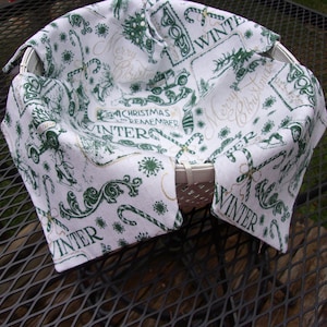 May include: A white wicker basket lined with a festive winter-themed fabric. The fabric features green and gold designs of candy canes, snowflakes, and the words "Merry Christmas" and "Winter". The basket is on a metal table.