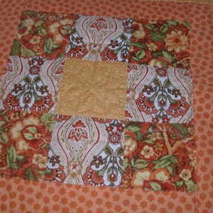May include: A square quilted fabric piece featuring a central solid yellow block surrounded by alternating patterned squares. The patterns include orange and green floral motifs and intricate paisley-like designs, all bordered by a small orange floral print.