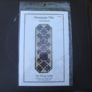 May include: A package containing a table runner pattern titled "Moroccan Tiles." The pattern features a geometric design in shades of red, brown, and beige. The package is sealed in clear plastic.