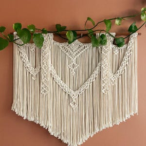 May include: Cream-colored macrame wall hanging with a geometric design and fringe. The wall decor is suspended from a wooden dowel and adorned with green leafy vines. This handcrafted piece adds a bohemian touch to any space.