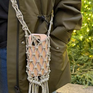 May include: A silver macrame phone holder with a pink phone inside. The holder has a woven design and a long strap for wearing around the neck. The holder is hanging on a person wearing a green jacket and blue jeans.