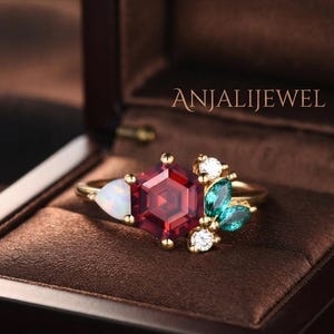 May include: A gold ring featuring a central hexagonal red garnet, a pear-shaped white opal, two marquise-cut green stones, and two small round clear stones. The text ANJALIJEWEL is visible in the background. The ring has a thin gold band.