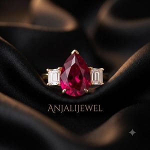 May include: A gold ring featuring a large pear-cut deep red gemstone in the center, flanked by two smaller emerald-cut clear stones on either side. The text ANJALIJEWEL is printed in a serif font below the ring, which rests on dark fabric.