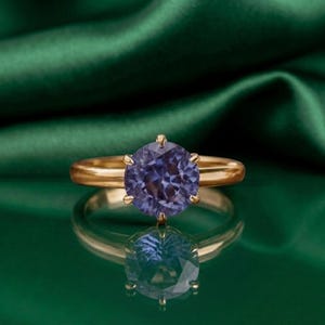 Può includere: Un anello in oro con una pietra preziosa rotonda e sfaccettata di colore viola-blu, fissata con una montatura a sei griffe. Il gambo è liscio e lucido, con una superficie riflettente che mette in risalto la chiarezza e il colore della pietra.