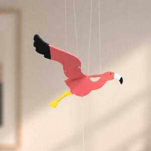 Könnte beinhalten: Ein hängendes Mobile in Form eines stilisierten rosa Flamingos mit schwarzen Flügelspitzen und einem gelben Bein. Der Flamingo ist an dünnen weißen Schnüren befestigt und hat ein kleines weißes Gewicht unter dem Körper für das Gleichgewicht.