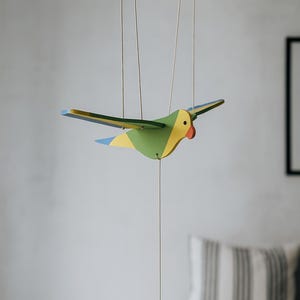 Può includere: Mobile a forma di uccello in legno colorato con le ali spiegate, sospeso con delle corde. L'uccello presenta dettagli dipinti in verde, giallo e blu, con un becco rosso. Un piccolo peso scuro è appeso sotto. Arredamento per la casa.