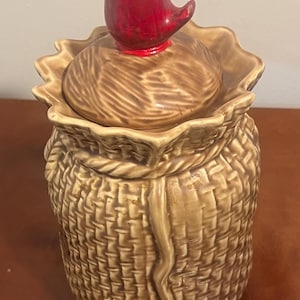 May include: A ceramic cookie jar shaped like a woven basket, with a lid topped by a red bird figurine. The jar is a warm brown color, with a detailed basket weave texture. The bird has a black eye and a red body.