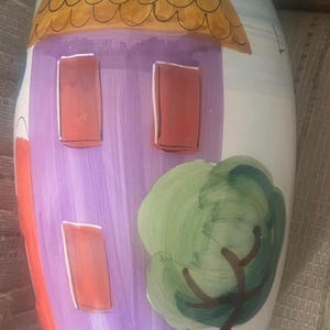 Op de afbeelding: Een handbeschilderde keramische vaas met een grillig huisontwerp. De vaas heeft een paars huis met rode ramen, een bruin dak en een groene boom. De bovenkant van de vaas is blauw en de algehele stijl is charmant en decoratief.