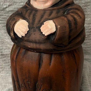 May include: A ceramic cookie jar in the shape of a monk. The jar is primarily brown with a light skin tone for the face and hands. The text "Salt Not Steel" is visible on the jar's base.