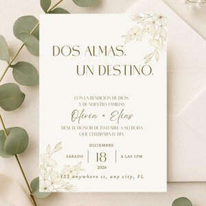 K&ouml;nnte beinhalten: Elfenbein-Hochzeitseinladung mit goldenen floralen Akzenten und Text. Die Einladung lautet "DOS ALMAS. UN DESTINO." und enth&auml;lt die Namen Olivia + Elias, Datum, Uhrzeit und Adresse. Ein passender Umschlag ist sichtbar.