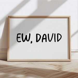 May include: A framed print with the text "EW, DAVID" in bold, black letters on a white background. The frame is a light brown color, and the print is displayed on a wooden surface.