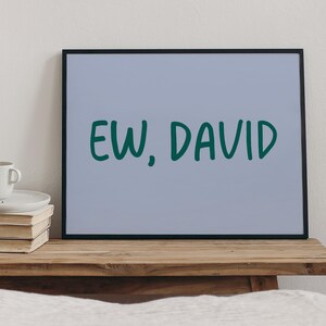 May include: A framed print with the phrase "EW, DAVID" in dark green lettering on a light blue background. The black frame sits on a wooden surface, with a stack of books and a mug visible to the left.