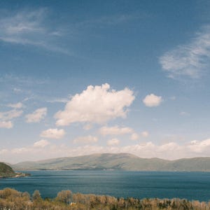 May include: A scenic landscape featuring a vast blue lake under a bright blue sky dotted with fluffy white clouds. Green hills and mountains frame the water, with a foreground of trees and vegetation.