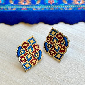 Può includere: Un paio di orecchini ornati con una base in metallo dorato. Ogni orecchino presenta un design simmetrico con dettagli in smalto rosso, blu e oro. Gli orecchini sono appoggiati su una superficie di legno chiaro con un tessuto blu sullo sfondo.