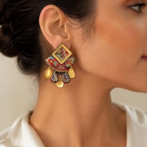 Puede incluir: Un pendiente llamativo con una pieza superior dorada en forma de diamante, una sección semicircular con un estampado floral en rojo, azul y oro, y cinco colgantes en forma de lágrima que alternan acabados en oro sólido y estampados.