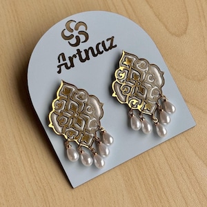 Puede incluir: Un par de pendientes ornamentados con un diseño de filigrana dorada, esmalte blanco y perlas de imitación colgantes. Los pendientes se exhiben en una tarjeta azul claro con el nombre de la marca "Artnaz" en escritura negra.