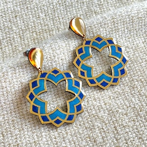 Op de afbeelding: Een paar goudkleurige oorbellen met een geometrisch ontwerp. De oorbellen hebben een gouden druppelvormige bovenkant en een cirkelvormige hanger met een gouden rand en blauwe en turquoise geometrische vormen. Op een getextureerd, gebroken wit oppervlak.
