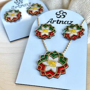 Può includere: Un set di gioielli con collana e orecchini abbinati. I pezzi presentano un design floreale con smalto rosso, bianco e verde, finiture color oro e un'emblema a forma di leone dorato. Il marchio "Artnaz" è visibile sulla scheda.