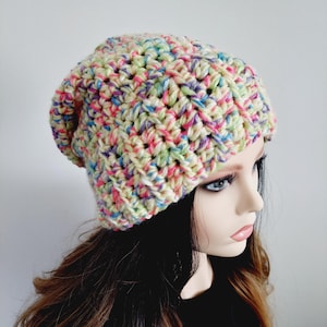 Op de afbeelding: Een handgemaakte gehaakte beanie met een gespikkeld patroon. De hoed heeft een mix van pastelkleuren, waaronder roze, blauw, groen en paars, op een crèmekleurige basis. De beanie is ontworpen om losjes en comfortabel te zijn.