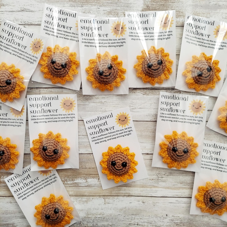Pu&ograve; includere: Spille a forma di girasole all'uncinetto con facce sorridenti, confezionate con biglietti. I biglietti riportano "emotional support sunflower". I girasoli sono gialli e marroni.