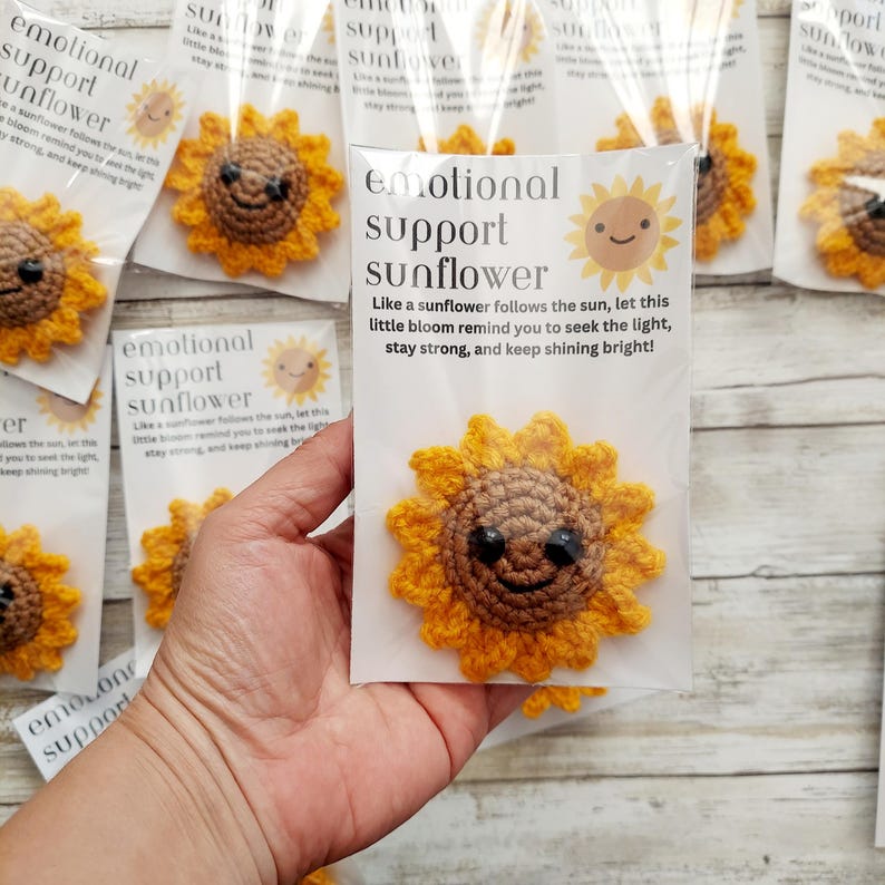 Pu&ograve; includere: Un girasole all'uncinetto con un volto sorridente &egrave; tenuto in una custodia trasparente. La confezione recita "emotional support sunflower" con una citazione sulla ricerca della luce e rimanere forti. Il girasole &egrave; giallo e marrone.