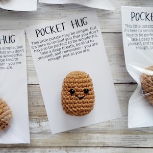 May include: A small, tan, crocheted potato figure with black bead eyes and a stitched smile, attached to a white card labeled POCKET HUG. The text reads: This little potato may be simple, but it's here to remind you that you don't have to be perfect to be wonderful. Take a deep breath, be kind to yourself, and remember - you are enough, just as you are.