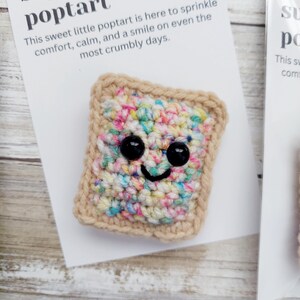 May include: A crocheted poptart with a smiling face and colorful sprinkles. The poptart is on a white card with the text "This sweet little poptart is here to sprinkle comfort, calm, and a smile on even the most crumbly days."