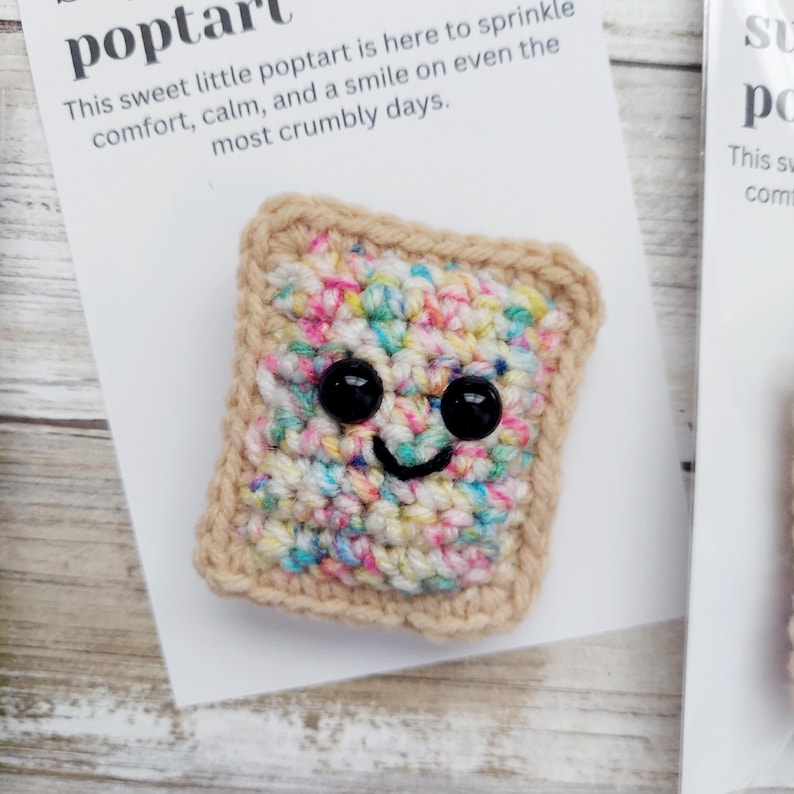 May include: A crocheted poptart with a smiling face and colorful sprinkles. The poptart is on a white card with the text "This sweet little poptart is here to sprinkle comfort, calm, and a smile on even the most crumbly days."