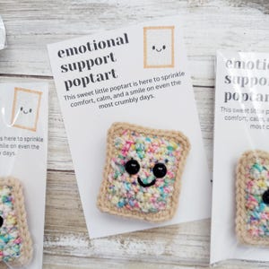 May include: A crocheted poptart with colorful sprinkles and a smiling face. The poptart is on a white card with the text "emotional support poptart" and "This sweet little poptart is here to sprinkle comfort, calm, and a smile on even the most crumbly days."