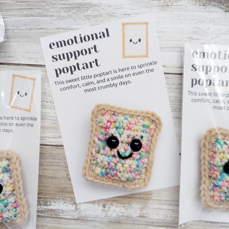 May include: A crocheted poptart with colorful sprinkles and a smiling face. The poptart is on a white card with the text "emotional support poptart" and "This sweet little poptart is here to sprinkle comfort, calm, and a smile on even the most crumbly days."