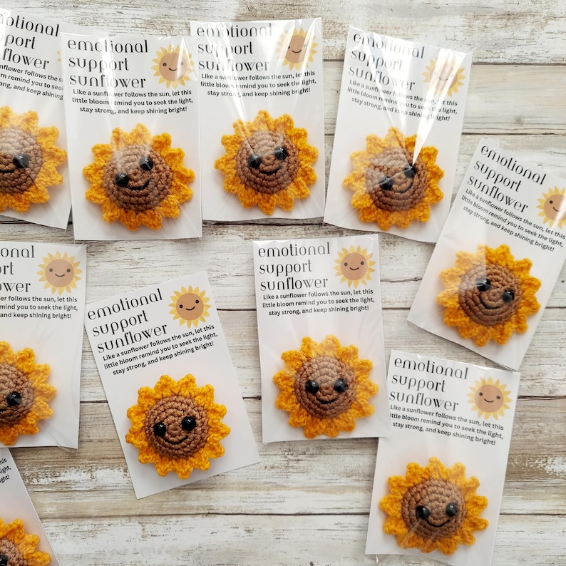 Pu&ograve; includere: Spille a forma di girasole all'uncinetto con facce sorridenti, confezionate con biglietti. I biglietti recitano "emotional support sunflower". I girasoli sono gialli e marroni. I biglietti hanno un sole.
