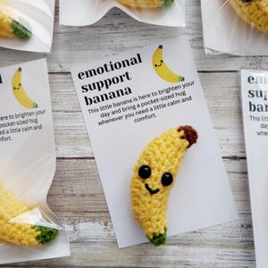 May include: Crocheted yellow banana with black eyes and a smiling mouth, packaged with a card that reads "emotional support banana." The card also states the banana is meant to brighten your day and bring comfort.