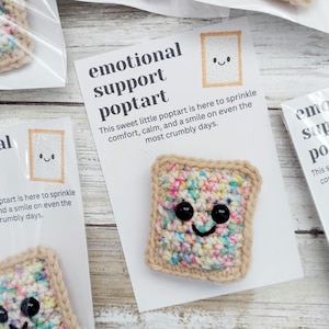 May include: A crocheted poptart with sprinkles and a smiling face. The poptart is on a white card with the text "emotional support poptart" and "This sweet little poptart is here to sprinkle comfort, calm, and a smile on even the most crumbly days."