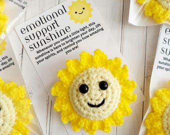 May include: Crocheted yellow sunflower with a smiling face, packaged with a card that reads "emotional support sunshine." The card includes text about brightening your day. Several of these items are visible.