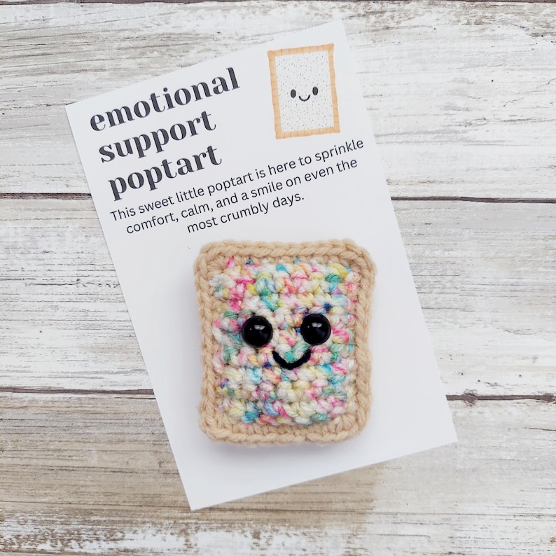 May include: A crocheted poptart with sprinkles and a smiling face. The poptart is on a white card with the text "emotional support poptart" and "This sweet little poptart is here to sprinkle comfort, calm, and a smile on even the most crumbly days."