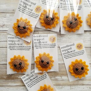 Pu&ograve; includere: Girasoli all'uncinetto con facce sorridenti, confezionati con biglietti. I biglietti recitano "emotional support sunflower" e presentano un'illustrazione di girasole. I girasoli sono gialli e marroni.
