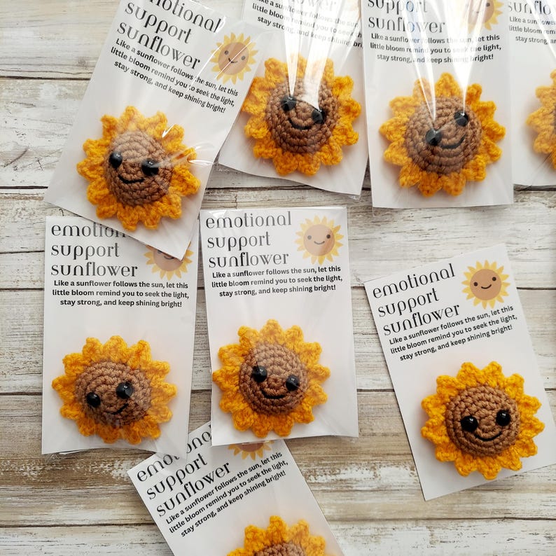 Pu&ograve; includere: Girasoli all'uncinetto con facce sorridenti, confezionati con biglietti. I biglietti recitano "emotional support sunflower" e presentano un'illustrazione di girasole. I girasoli sono gialli e marroni.