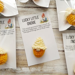 May include: Crocheted gnome with an orange body and a cream-colored hat, displayed on a white card. The card reads "LUCKY LITTLE GNOME" and includes text about luck and happiness. Several gnomes are visible.