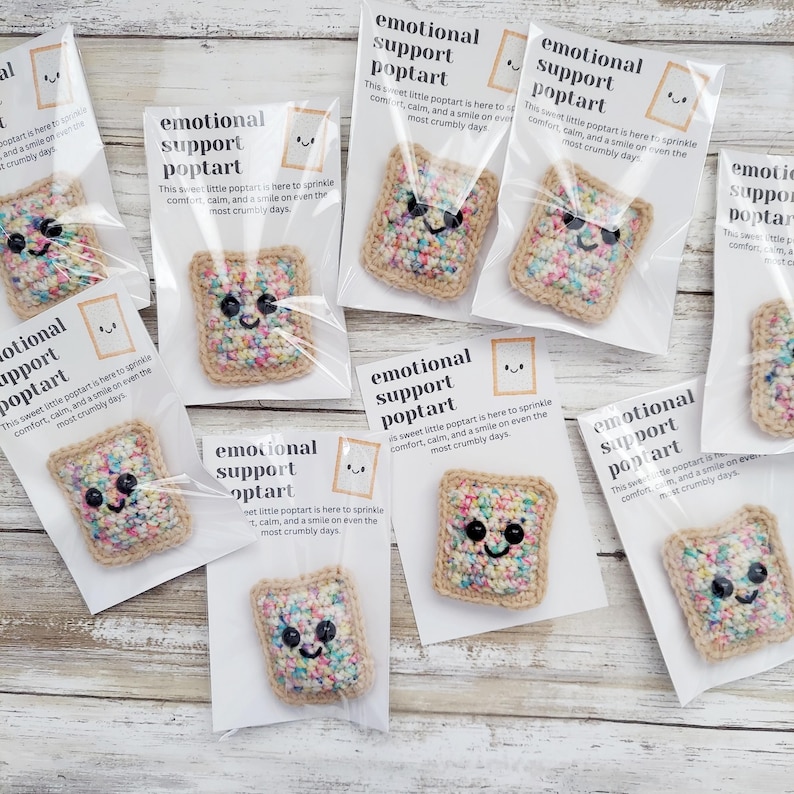 May include: A collection of nine small crocheted poptart-shaped plushies with colorful sprinkles. Each poptart has a smiling face and is packaged in a clear plastic bag with a white card that reads "Emotional Support Poptart. This sweet little poptart is here to sprinkle comfort, calm, and a smile on even the most crumbly days."