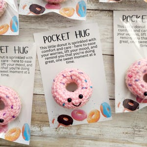 May include: Pink crocheted donut with a smiling face, packaged with a card that reads "Pocket Hug." The card also features watercolor illustrations of donuts. The donut is sprinkled with blue and purple yarn.