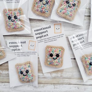 May include: Four crocheted poptart pins with colorful sprinkles and smiling faces. Each pin is packaged in a clear plastic bag with a white card that reads "emotional support poptart" and a short message about the poptart being there to sprinkle comfort, calm, and a smile on even the most crumbly days.