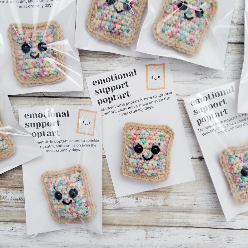 May include: Four crocheted poptart pins with colorful sprinkles and smiling faces. Each pin is packaged in a clear plastic bag with a white card that reads "emotional support poptart" and a short message about the poptart being there to sprinkle comfort, calm, and a smile on even the most crumbly days.