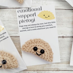 May include: A crocheted pierogi with black button eyes and a smiling mouth, presented on a card that reads "emotional support pierogi." The card also includes the text "Hugs may not always be near, but this little pierogi is here to bring you comfort, warmth, and a smile whenever you need it!"