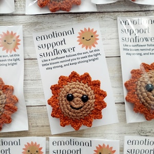 May include: A crocheted sunflower with a tan center, orange petals, and black plastic eyes and a stitched smile. It is attached to a white card with the text "emotional support sunflower" and a poem about seeking the light and staying strong.