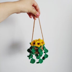 May include: A small crocheted hanging planter with a brown pot and green leaves. The planter is filled with yellow crocheted sunflowers.