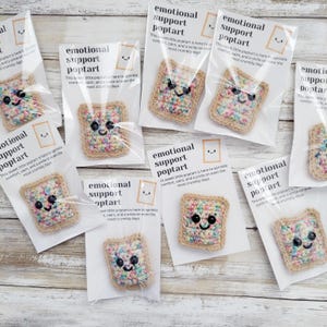 May include: A collection of nine small crocheted poptart-shaped plushies with colorful sprinkles. Each poptart has a smiling face and is packaged in a clear plastic bag with a white card that reads "Emotional Support Poptart. This sweet little poptart is here to sprinkle comfort, calm, and a smile on even the most crumbly days."