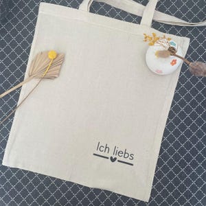 Könnte beinhalten: Beige Canvas-Tragetasche mit langen Henkeln. Der Beutel zeigt den Schriftzug "Ich liebs" in schwarzer Schrift mit einem Herzsymbol. Dekorative Trockenblumen und eine kleine Vase sind auf der Tasche platziert.