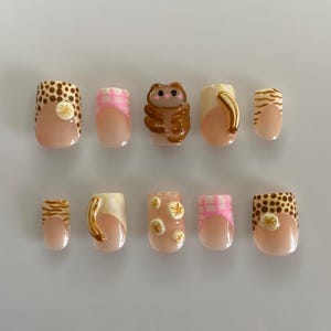 May include: A set of ten artificial nails with diverse designs. The nails showcase patterns such as polka dots, stripes, and floral details. Some nails have a 3D bear design, and others have a coffee cup design. The nails are in beige, pink, and brown hues.